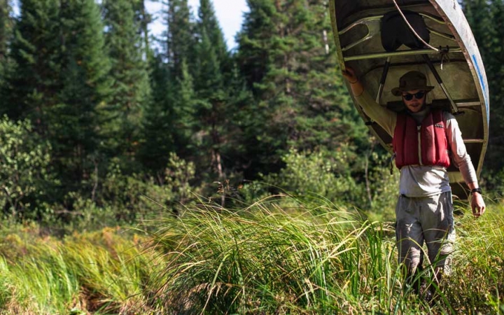 a young person wearing a life jacket carries a canoe on their shoulders through tall green grass